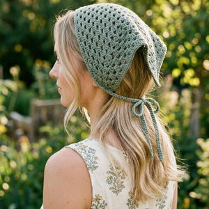 May include: A sage green crochet headscarf with a square shape and tie closure. The head covering is made of a textured, open-weave fabric. The scarf is tied at the nape of the neck with a bow. The background is a blurred outdoor scene.
