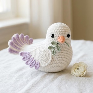 May include: A handmade, crocheted white dove with lavender and green accents. The dove has a peach-colored beak and holds a green sprig in its beak. A white flower rests beside the dove on a white surface.