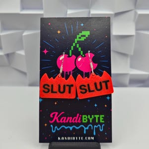 May include: A pair of novelty earrings featuring a cherry design with the word "SLUT" in black on a red background. The earrings hang from silver hooks and are displayed on a black card with the "KandiBYTE" logo and website.