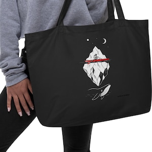 Puede incluir: Bolso tote negro con un gráfico blanco y rojo de un iceberg, una persona en un bote, una ballena y elementos celestiales. El bolso tiene asas negras y lo sostiene una persona.
