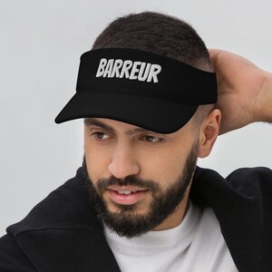 May include: Black visor with the word "BARREUR" in white. A person is wearing the visor, a white t-shirt, and a black sweater draped over the shoulders.