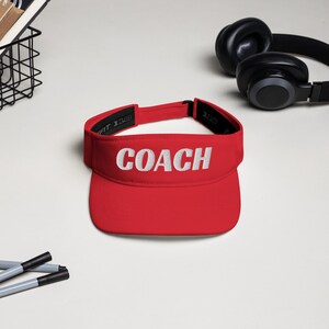 Puede incluir: Una visera roja brillante con la palabra "COACH" bordada en blanco en la parte delantera. La visera está hecha de un material ligero y tiene una visera curvada. Auriculares negros en el fondo.