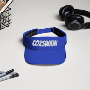 May include: A vibrant blue visor featuring the white embroidered word "COXSWAIN". The visor has a curved peak and an adjustable strap. Black headphones are visible in the background.