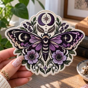 May include: An embroidered patch featuring a central moth with purple and black wings, adorned with crescent moon and star motifs. The design includes lavender sprigs, purple flowers, and white lily of the valley, all set against a cream background.