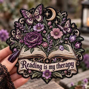 May include: An embroidered patch featuring an open book with purple flowers and butterflies emerging from the pages. A crescent moon and stars are at the top. A banner at the bottom contains the text Reading is my therapy in purple.