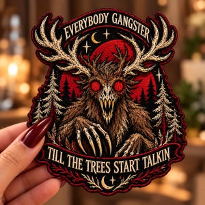 May include: An embroidered patch featuring a central creature with large antlers, glowing red eyes, and sharp teeth. The background shows a red moon and pine trees. Text reads: EVERYBODY GANGSTER TILL THE TREES START TALKIN. The design uses black, red, and cream thread.
