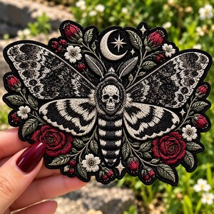 May include: An embroidered patch featuring a central moth with a skull pattern on its thorax, surrounded by red roses, white flowers, and green leaves. A white crescent moon and a four-pointed star are positioned above the moth's head.