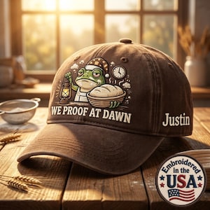 May include: A brown baseball cap featuring an embroidered graphic of a frog wearing a nightcap, holding a lantern, and standing next to a basket of dough. The text WE PROOF AT DAWN is printed below the graphic, with the name Justin on the side.