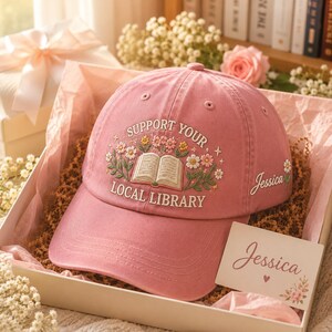 May include: A pink baseball cap with the embroidered text "Support Your Local Library" and a floral design. The cap is in a gift box with a card that says "Jessica".