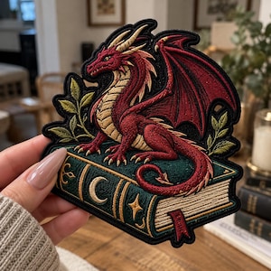 May include: An embroidered patch featuring a red dragon with gold accents perched atop a dark green book. The book cover displays a gold crescent moon and star symbol. Green leafy branches frame the dragon on both sides of the patch.