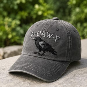 May include: A charcoal gray baseball cap with a distressed finish, featuring an embroidered black crow graphic and the white text F-CAW-F arched above it. The cap has a curved brim, stitched eyelets, and a structured crown with a top button.
