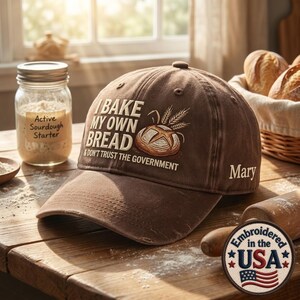 May include: A brown baseball cap featuring the embroidered text I BAKE MY OWN BREAD & DON'T TRUST THE GOVERNMENT, a graphic of a loaf of bread with wheat stalks, and the name Mary on the side. A circular patch reads Embroidered in the USA.