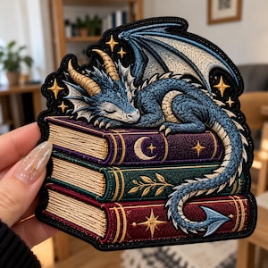 May include: An embroidered patch featuring a blue dragon with gold horns sleeping atop a stack of three books. The top book is purple with a moon and star, the middle is green with leaf motifs, and the bottom is red with a starburst design.