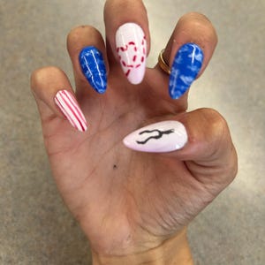 May include: A hand with long, almond-shaped nails, each painted with a unique design. The nails feature stripes, baseball stitching, and blue marble effects. One nail has a black curved design on a white base.