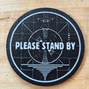 Puede incluir: Un posavasos redondo negro con borde blanco. Presenta el texto "PLEASE STAND BY" y un gráfico de patrón de prueba de televisión retro, incluyendo una figura estilizada. Adecuado para diversas bebidas.