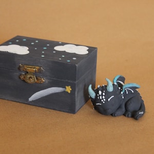 May include: A dark gray wooden box with a brass clasp, painted with white clouds, stars, and a shooting star. Beside it is a black figurine of a dragon with blue horns and wings.