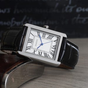 May include: A rectangular silver wristwatch with a white face, black Roman numerals, and blue hands. The watch has a black leather strap with a crocodile pattern. The brand name "ERIKSEN" is printed on the watch face.