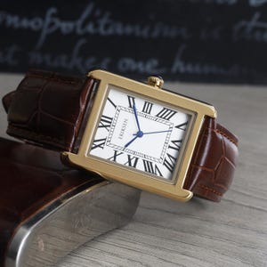 May include: A gold rectangular wristwatch with a white face, black Roman numerals, and blue hands. The watch has a brown leather strap with a crocodile pattern. The watch face reads "ERIKSEN".