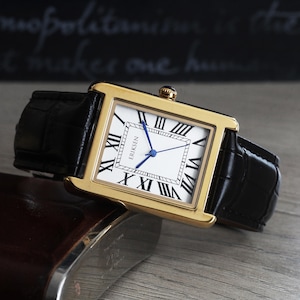 May include: A gold rectangular wristwatch with a white face, black Roman numerals, and blue hands. The watch has a black leather strap and the brand name "Eriksen" is visible on the watch face. The watch is resting on a wooden surface.