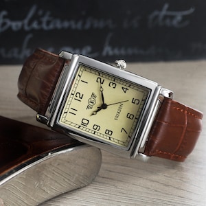 May include: A rectangular wristwatch with a silver-tone case and a brown leather strap featuring a crocodile-embossed texture. The watch face is cream-coloured with black Arabic numerals, black hands, and the text ERIKSEN with a winged E logo.