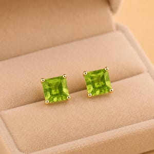 Peut inclure: Une paire de boucles d'oreilles à clous avec des péridots de taille carrée montés sur métal doré. Chaque boucle présente une pierre précieuse vert lime vibrante à facettes, fixée par quatre petites griffes dorées aux coins.