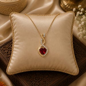 Può includere: Una collana in oro con un ciondolo caratterizzato da una pietra preziosa rossa a forma di cuore al centro, circondata da piccole pietre trasparenti. Il ciondolo ha un design intrecciato che si estende verso l'alto, decorato con pietre chiare.