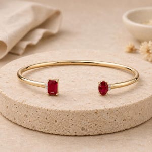Puede incluir: Una pulsera abierta de oro con un rubí rojo de corte rectangular en un extremo y un rubí rojo de corte ovalado en el otro. Cada rubí está flanqueado por dos pequeñas piedras de acento redondas y transparentes engastadas en garras de oro.