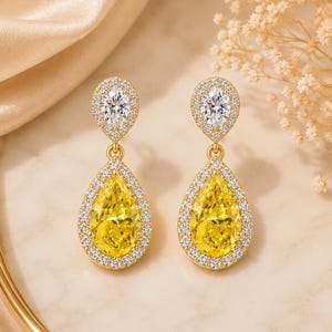 Peut inclure: Une paire de boucles d'oreilles pendantes en or avec une pierre jaune en forme de poire entourée d'un halo de petites pierres claires. Au-dessus, une pierre claire ovale plus petite est également entourée d'un halo de pierres claires.