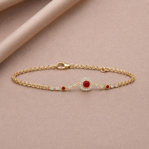 Può includere: Un bracciale a catena dorata con una pietra rossa circolare centrale circondata da un alone di piccole pietre chiare, affiancato da due pietre rosse più piccole e ulteriori pietre chiare lungo la sottile catena a maglie con chiusura.
