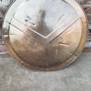 May include: A bronze-coloured shield with a distinctive V-shaped pattern in the centre. The shield is circular with a slightly textured surface. The background features a brick wall and a concrete surface.