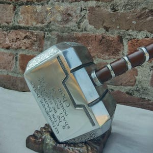 May include: A silver and brown replica of Thor's hammer, Mjolnir, resting on a brown stand. The hammer head has engraved text and decorative patterns. The handle is wrapped in brown leather with silver accents. The text on the hammer reads "WHOSOEVER HOLDS THIS HAMMER IF HE BE WORTHY SHALL POSSESS THE POWER OF THOR."