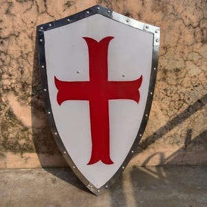 Könnte beinhalten: Ein Schild im mittelalterlichen Stil mit weißem Hintergrund und einem großen roten Kreuz in der Mitte. Der Schild hat einen silbernen Metallrand mit Nieten. Ein historisches oder dekoratives Objekt.