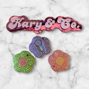 May include: A collection of bedazzled flower-shaped phone grips in green, purple, and pink. The grips have a circular button for easy use. The image also features a pink and red mirrored sign that reads "Kary & Co."