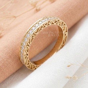 May include: A gold ring featuring a decorative filigree pattern and a line of small, clear stones. The ring has the text "Smiling Stone Jewels" engraved on the side. The ring is displayed against a soft, neutral backdrop.
