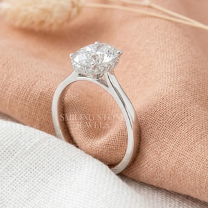 May include: A silver engagement ring with a large, oval-cut diamond center stone and a halo of smaller diamonds. The ring is set on a delicate band. The text "SMILING STONE JEWELS" is visible on the ring.