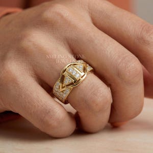 May include: A gold ring with a geometric design, featuring a central triangle and smaller triangles, all encrusted with small, sparkling stones. The ring is on a finger, with the text "SMILING STONE JEWEL" visible.
