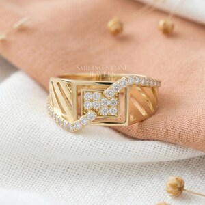 May include: A gold-colored ring with a geometric design. The ring features a square center with small, clear stones and a band of clear stones. The ring has a rectangular shape with cut-out details. The text "SMILING STONE JEWELS" is visible.