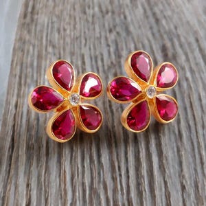 May include: A pair of gold-coloured flower-shaped stud earrings. Each earring features five teardrop-shaped, deep red gemstones surrounding a small, clear gemstone in the centre. The earrings are set against a wooden background.