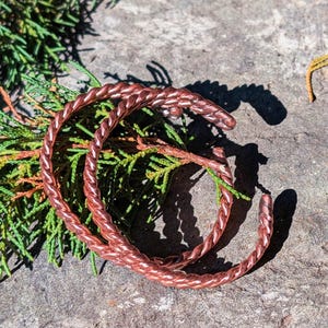 Op de afbeelding: Twee koperkleurige oorringen van gedraaid metaal liggen op een stenen oppervlak met groene bladeren. De oorbellen hebben een gevlochten touwontwerp en zijn aan de onderkant open. De oorbellen zijn warm roodbruin.