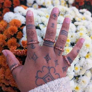 May include: A hand adorned with five copper-colored braided rings. The hand is covered in tattoos and is set against a backdrop of orange and white flowers. The rings are a simple, yet elegant design.