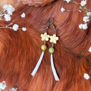 May include: A pair of dangle earrings featuring cream-colored flower-shaped beads, green beads, and white tassel-like accents. The earrings are suspended from bronze-colored ear wires. The background is a reddish-brown fur-like texture, with white flowers.