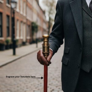 May include: A vintage-style walking cane with a brass handle and a dark red wooden shaft. The cane is held by a person wearing a dark gray suit. The text "Engrave your Text/date here" is visible.