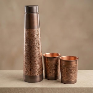 May include: A copper water bottle and two matching cups. The bottle has a dark brown cap and base, with an ornate floral pattern etched into the copper. The cups also feature the same etched design. The set is displayed on a neutral surface.
