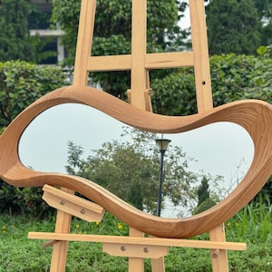 May include: A wall mirror featuring an irregular, organic, amoeba-like shape with a thick, natural light brown wood grain frame. The mirror is mounted on a light-colored wooden easel, displaying a reflective surface with a visible wood texture.