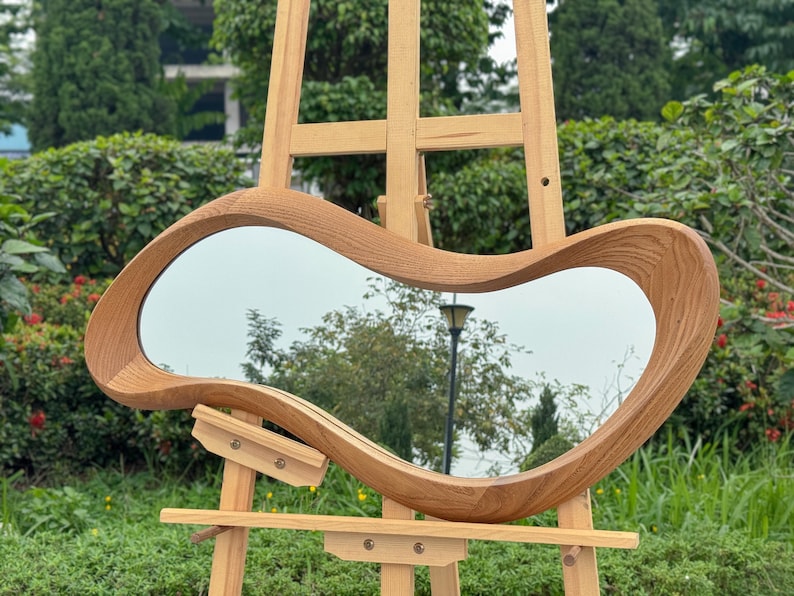 May include: A wall mirror featuring an irregular, organic, amoeba-like shape with a thick, natural light brown wood grain frame. The mirror is mounted on a light-colored wooden easel, displaying a reflective surface with a visible wood texture.