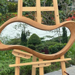 May include: An irregular, organic-shaped mirror featuring a thick, light-toned wooden frame with a visible grain pattern. The mirror is mounted on a light-colored wooden easel stand, reflecting a garden scene with green trees and shrubs.