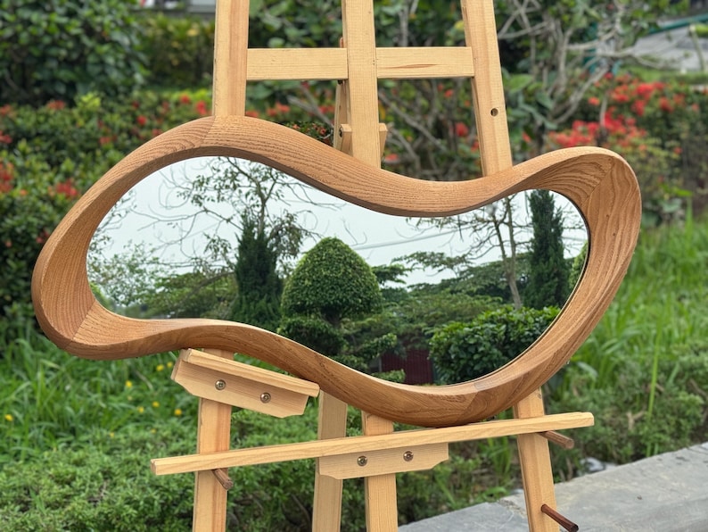 May include: An irregular, organic-shaped mirror featuring a thick, light-toned wooden frame with a visible grain pattern. The mirror is mounted on a light-colored wooden easel stand, reflecting a garden scene with green trees and shrubs.