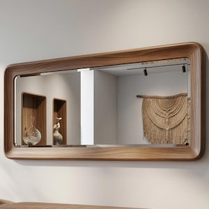 May include: A rectangular wooden-framed mirror with rounded corners hangs on a white wall. The mirror reflects a room with decorative shelves, vases, and a macrame wall hanging. The wood frame has a natural grain.