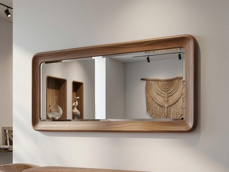 May include: A rectangular wooden-framed mirror with rounded corners hangs on a white wall. The mirror reflects a room with decorative shelves, vases, and a macrame wall hanging. The wood frame has a natural grain.