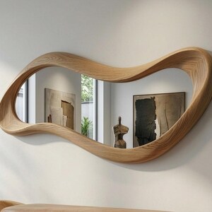 May include: A wall-mounted mirror featuring an irregular, wavy, organic-shaped frame crafted from light-toned wood with a visible grain pattern. The reflective surface displays a room interior with a framed painting and a small sculpture.